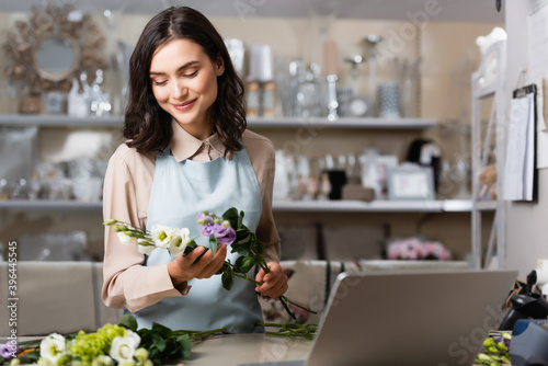 smiling florist holding eustoma flowers near laptop on blurred foreground