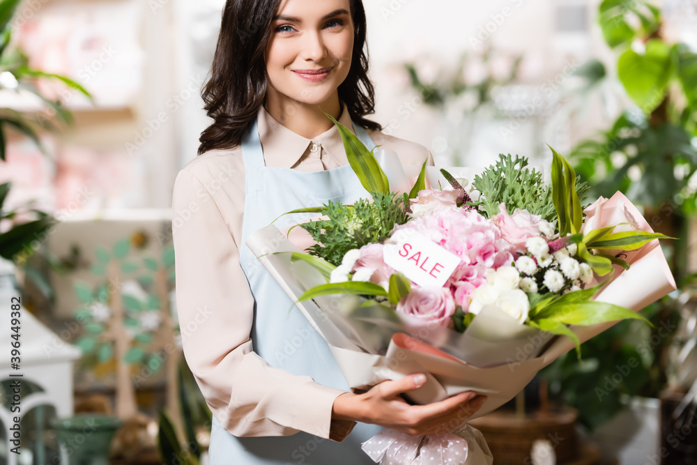 happy florist holding bouquet with sale lettering on tag while looking at camera near blurred plants on background happy florist holding bouquet with sale lettering on tag while looking at camera near blurred plants on background