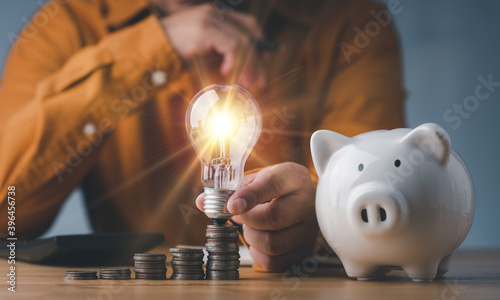 saving energy and money concept. idea for save or investment. businessman holding lightbulb on piggy bank and coins stacking on desk with calculator.