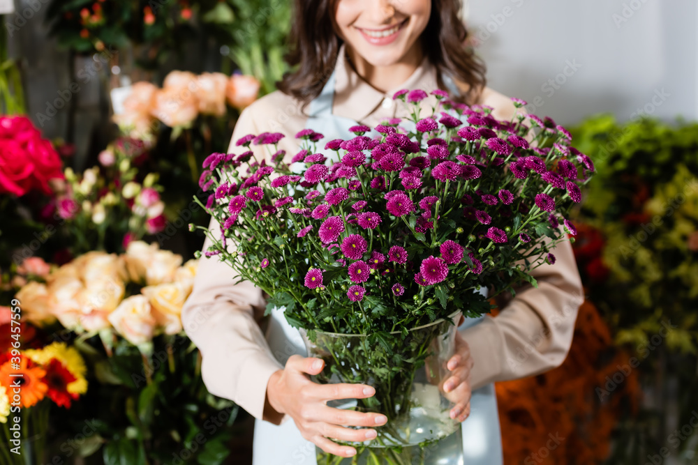Cropped view of female florist holding vase with purple chrysanthemums near blurred range of flowers on background