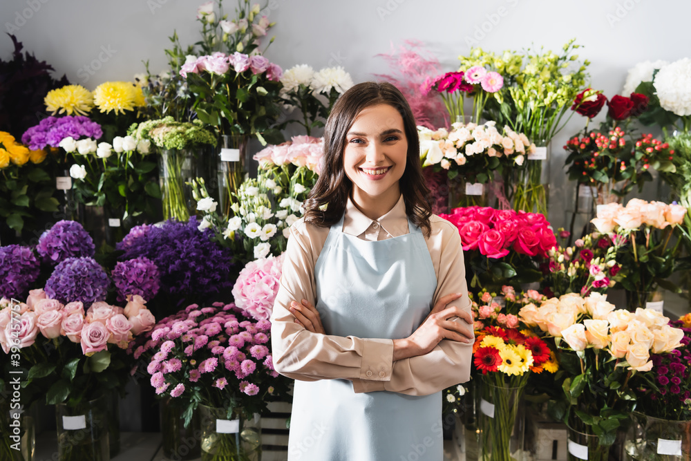 Smiling female florist with crossed arms looking at camera, while standing near racks of flowers on background