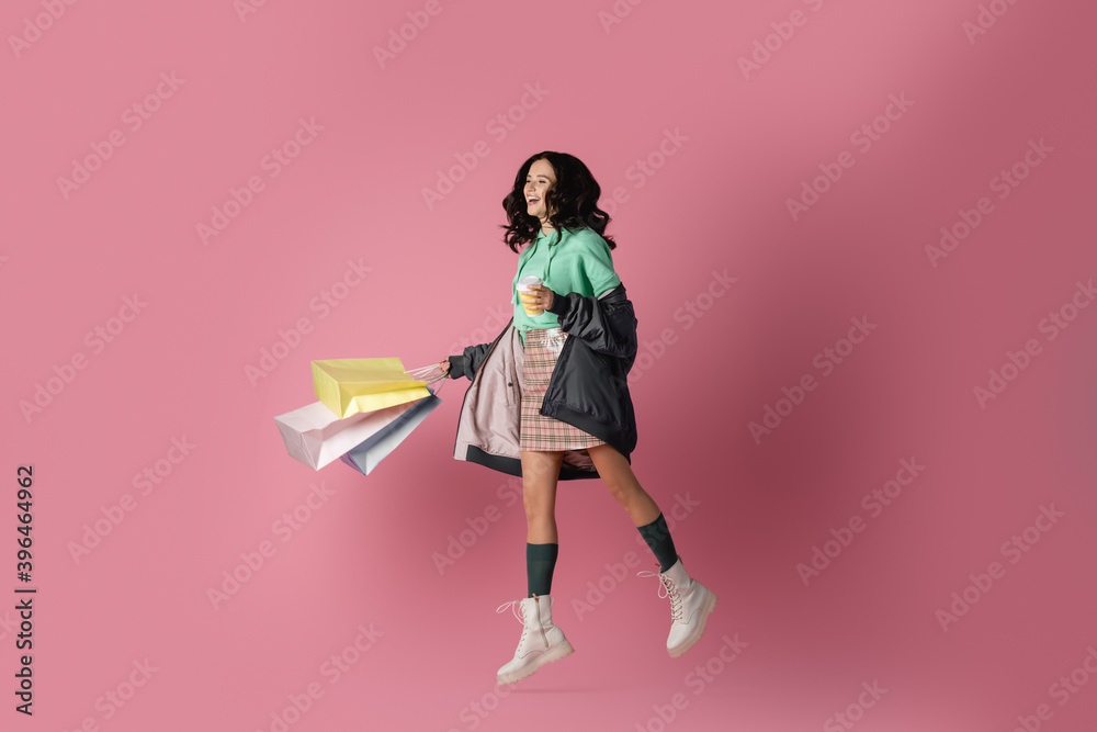 brunette young woman in casual winter outfit with shopping bags and coffee to go jumping on pink background brunette young woman in casual winter outfit with shopping bags and coffee to go jumping on pink background