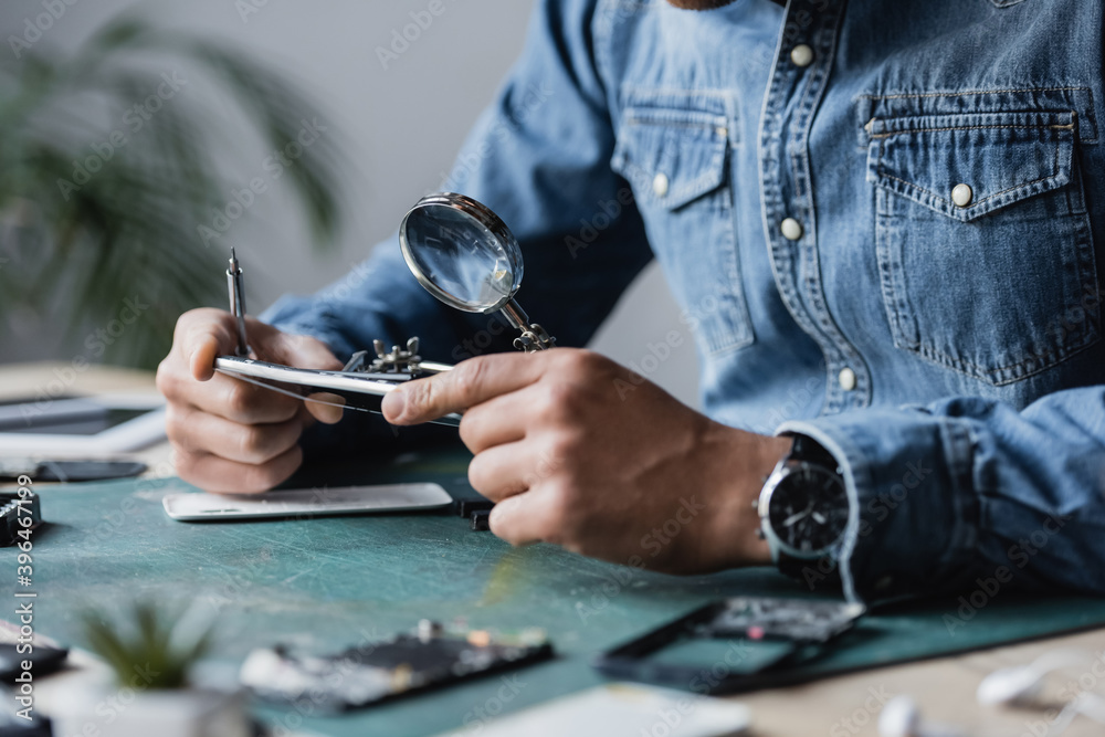 Cropped view of repairman with screwdriver holding disassembled cellphone near magnifier at table on blurred foreground