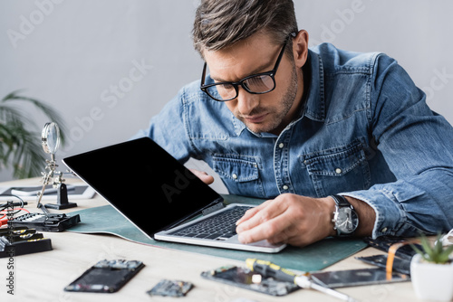 Focused repairman with broken key looking at keyboard of damaged laptop at workplace on blurred foreground