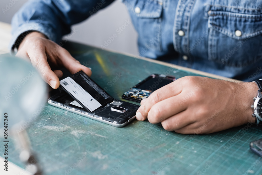 Cropped view of repairman putting battery in mobile phone on table with blurred magnifier on foreground Cropped view of repairman putting battery in mobile phone on table with blurred magnifier on foreground