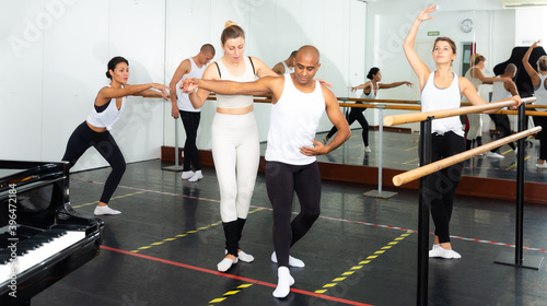 European female ballet teacher showing moves in front of a group of multiracial ..dancers in large dancing studio
