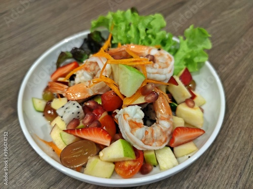 Mixed seasonal fruit spicy salad with shrimp in white plate on wooden table.