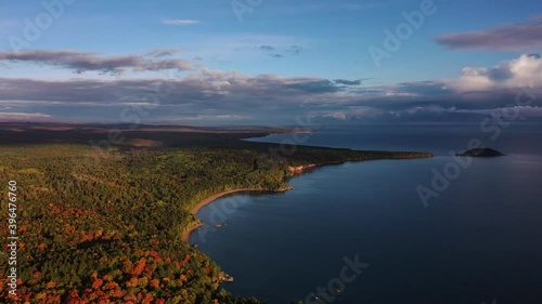 Beautiful aerial panning shot over the coastline of Lake Superior with tree covered forests below and clouds in the blue sky above.