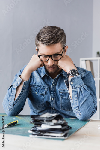 Thoughtful repairman with hands near cheeks sitting at table wit blurred pile of broken mobile phones on foreground