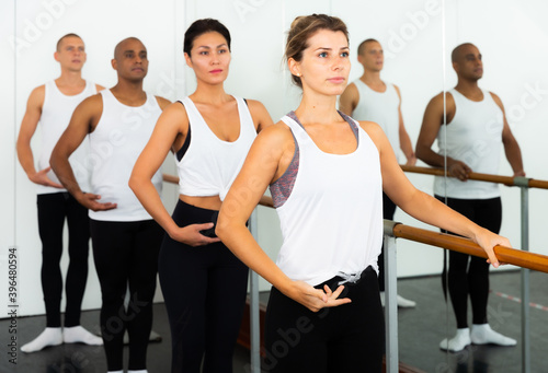 Group of people doing ballet exercises using barre in gym with focus to fit athletic toned woman in foreground in health and fitness concept