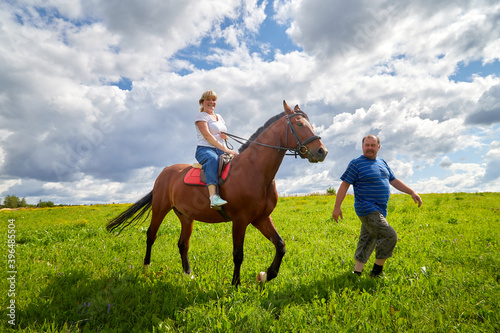 Kirov, Russia - August 08, 2020: Girl riding on a horse and fat man who is groom walking near them on green field and blue sky with white clouds on background