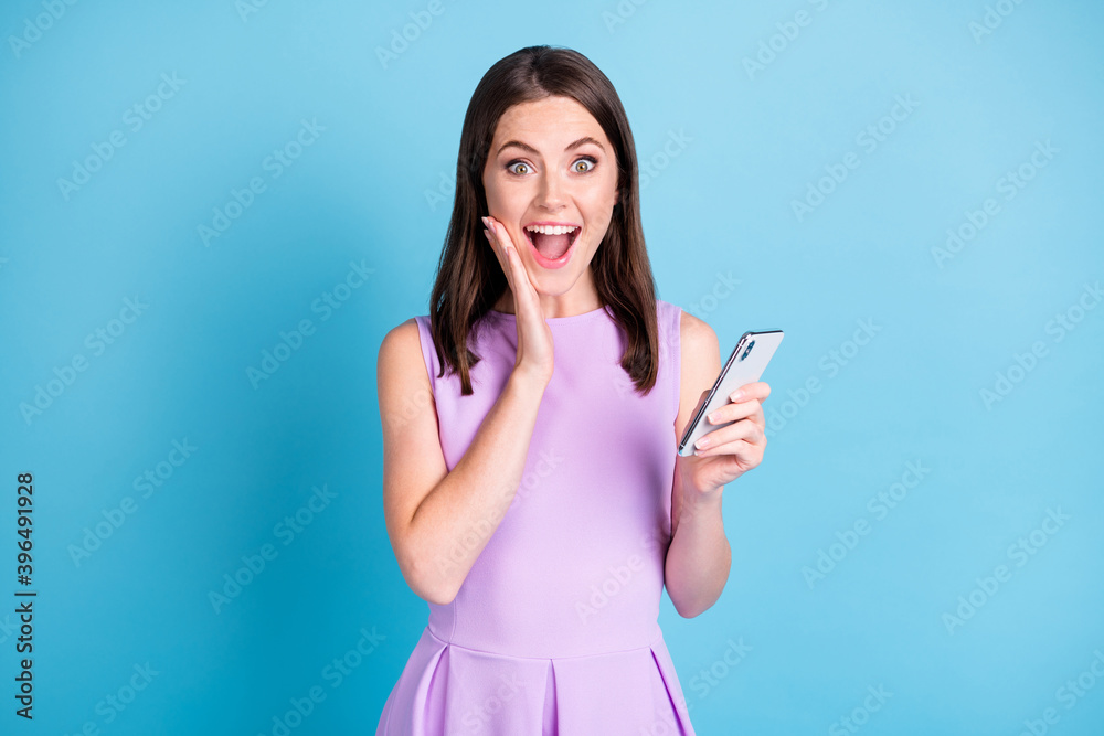 Photo of shocked happy young lady hold phone hand cheekbone amazed isolated on blue color background Photo of shocked happy young lady hold phone hand cheekbone amazed isolated on blue color background