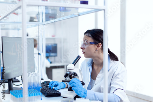 Laboratory technician working in microbiology lab using computer and microscope. Chemist wearing lab coat using modern technology during scientific experiment in sterile environment.