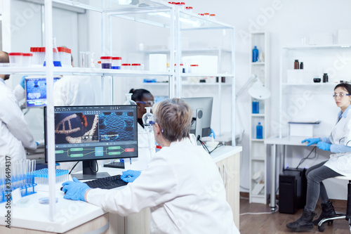Elderly virologist working on computer with virus on display in modern facility. Senior scientist in pharmaceuticals laboratory doing genetic research wearing lab coat with team in the background.