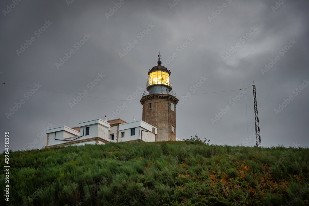 old lighthouse on a coastal hill of a small town, the lighthouse has a yellow flashing light, it is night and the sky is dramatic, gray with clouds