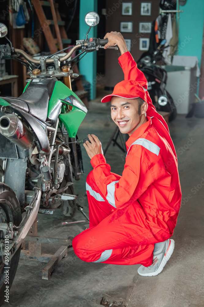 male mechanic smiles while setting up the carburetor using the screwdriver next to the motorbike in the garage