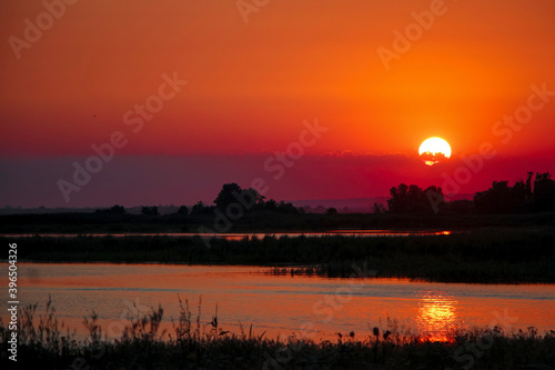 Beautiful red and orange sunset over the river. Golden twillight  sunlight reflected in ripples of water. Dark silhouette of trees.