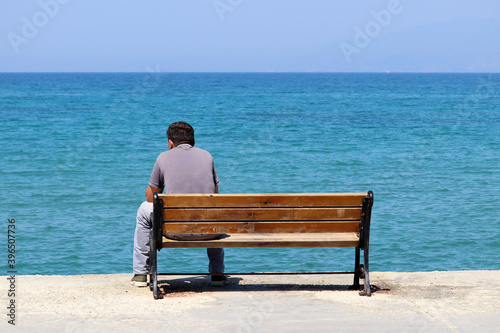 Lonely man sitting on a wooden bench on blue sea background, rear view. Vacation on a beach during coronavirus pandemic