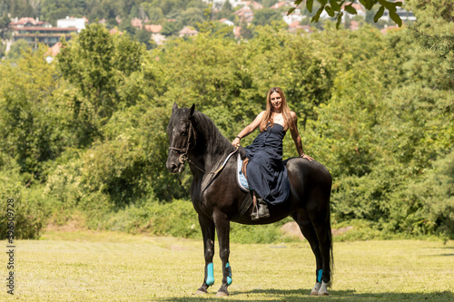 Girl with a long copper hair and long flowy shoulderless dress, ride a black horse with trees in the background.