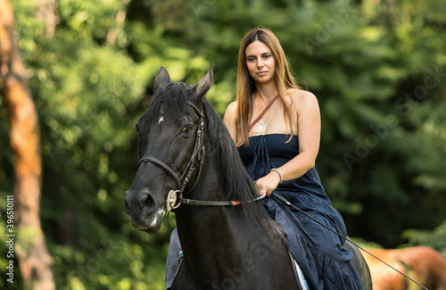Girl with a long copper hair and long flowy shoulderless dress,  ride a black horse with trees in the background. © Deya