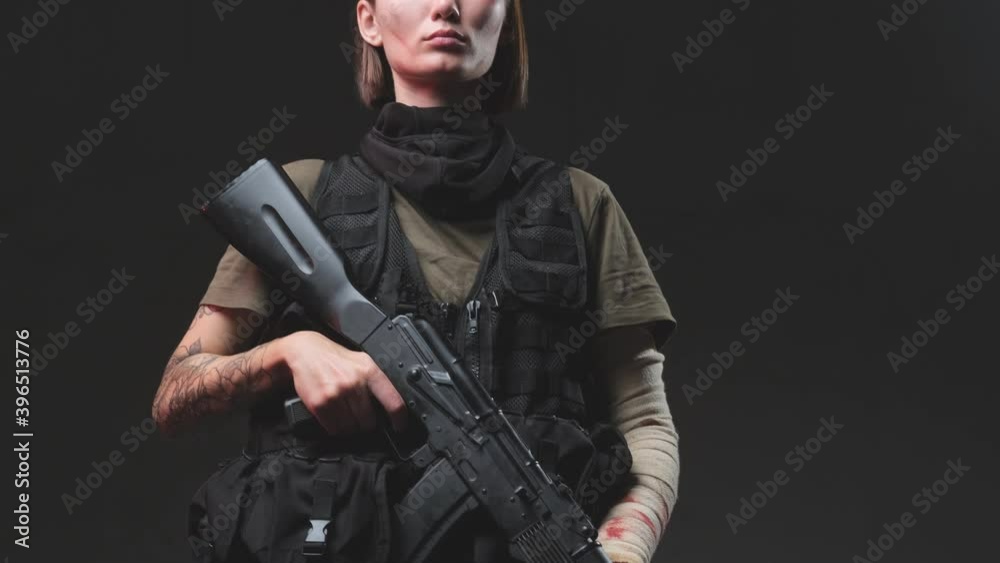Posing in dark background beautiful female mercenary dressed in dark martial clothing and holding ak 74 rifle with tattooed and bandaged hands. Posing in dark background beautiful female mercenary dressed in dark martial clothing and holding ak 74 rifle with tattooed and bandaged hands.