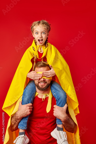 shocked super girl sits on neck of father closing his eyes, superhero man hold child and smiles. kid girl with opened mouth looks at camera isolated over red background