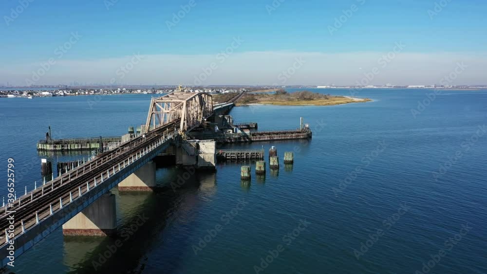 An aerial shot over Grassy Bay in Queens, NY. The camera dolly in along the elevated train tracks with a swing bridge over the bay. It is a peaceful scene shot on a beautiful day with blue skies.