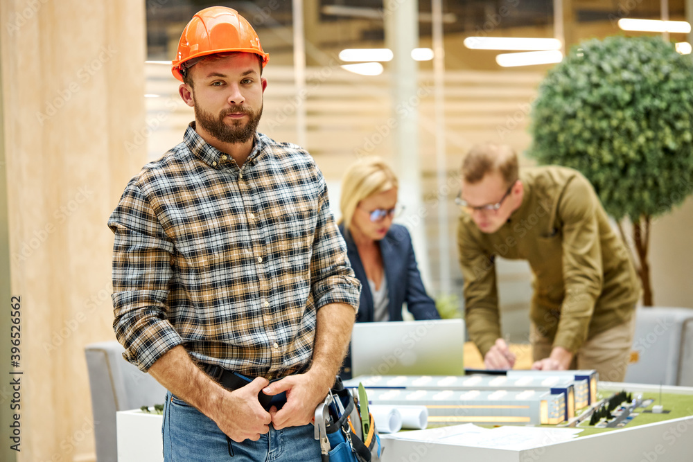 portrait of architect man in helmet in the office with talking collagues engineers in the background, he looks at camera posing portrait of architect man in helmet in the office with talking collagues engineers in the background, he looks at camera posing