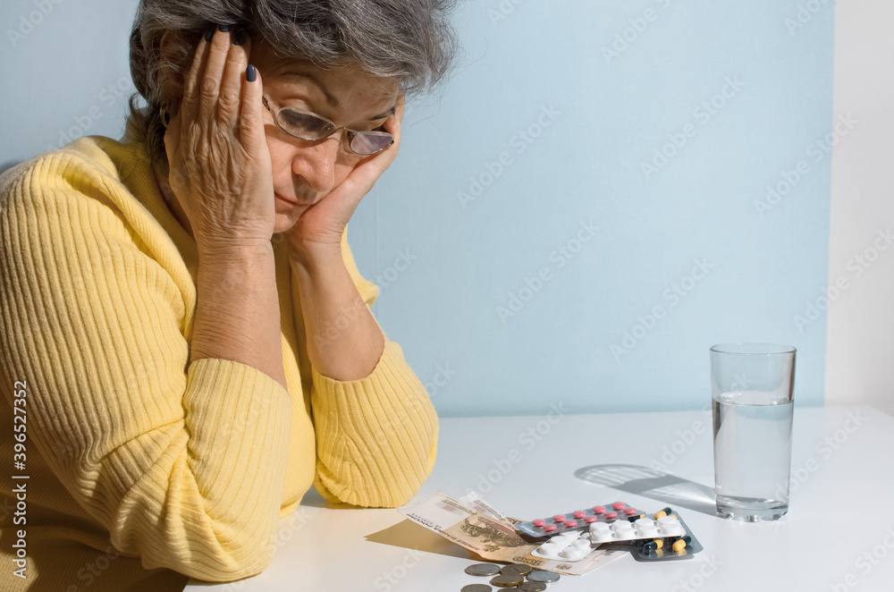 Fototapeta premium Elderly woman with glasses looking at money and pills on the table. Concept of depression, drug price, treatment cost