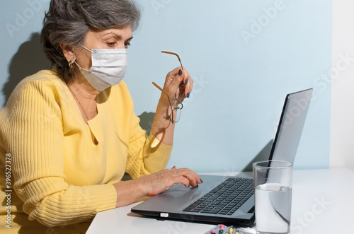 Elderly woman with glasses and medical mask using laptop at home. Concept for online consultation with doctor, internet search, self-isolation, new normal.