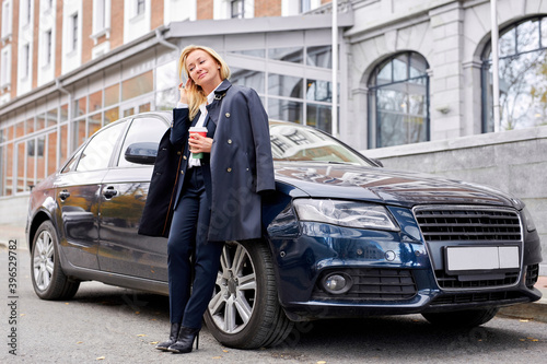 business woman posing next to her luxurious car outdoors in city, stand holding cup of coffee © Roman