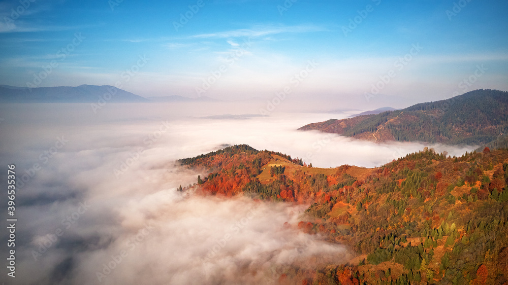 Beautiful Autumn mountain panorama. Morning thick fog cover valley. Sunny landscape with meadow and colorful forest