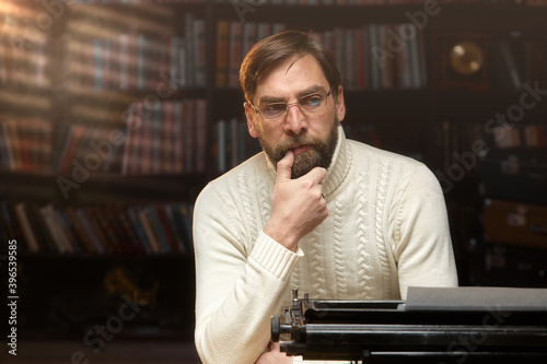A focused male writer with glasses is typing a novel on a typewriter. A man with a beard looks at the camera, sitting at the home table, in the background of the library