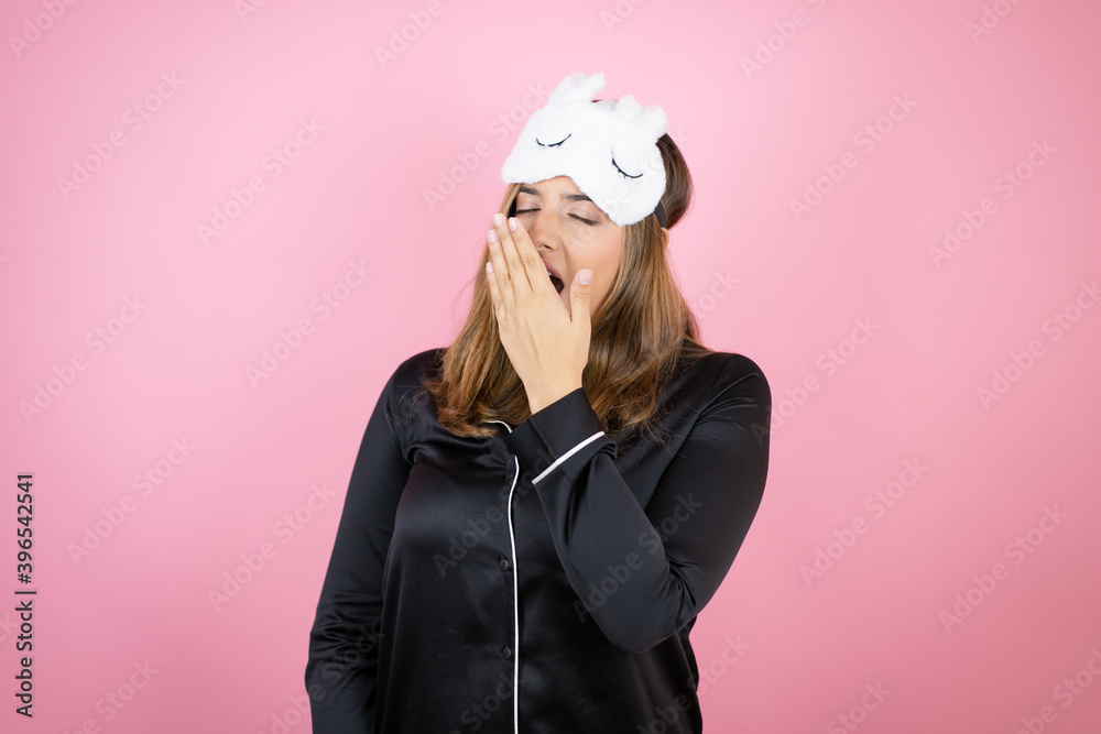 Young caucasian woman wearing sleep mask and pajamas over isolated pink background bored yawning tired covering mouth with hand. Restless and sleepiness.