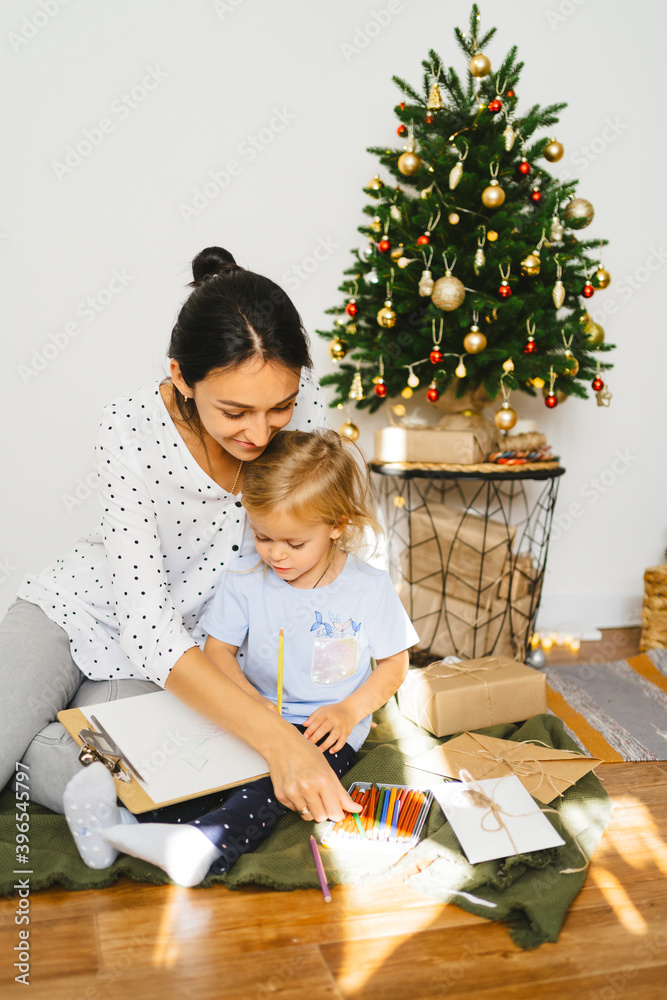 .Mom and daughter are writing a letter to santa claus on the background of a christmas tree and gifts. Young woman teaches little cute girl to draw a New Year card. Family cozy moments.. .Mom and daughter are writing a letter to santa claus on the background of a christmas tree and gifts. Young woman teaches little cute girl to draw a New Year card. Family cozy moments..