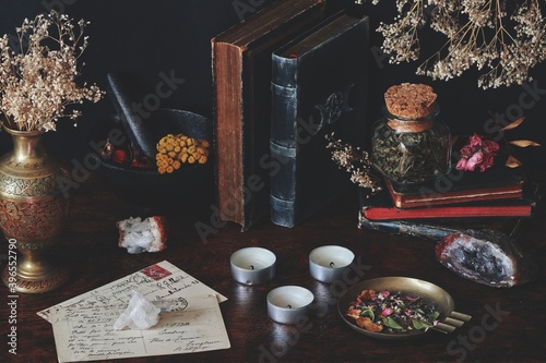 A collection of old vintage books on wiccan witch altar. Worn books placed on black table, with nature themed items on it, like dried herbs, flowers, crystals, old postcards, mortar vase white candles
