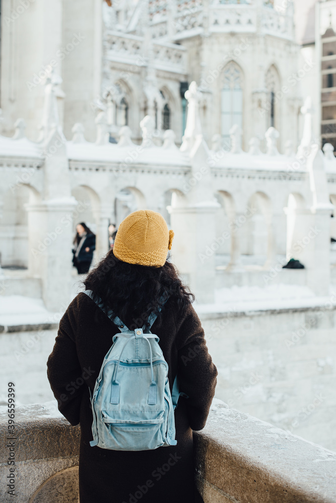 Back view of unrecognisable young woman tourist in winter coat and yellow hat with blue backpack looking at gothic architecture of Fisherman Bastion in Budapest, Hungary during bright winter day in De