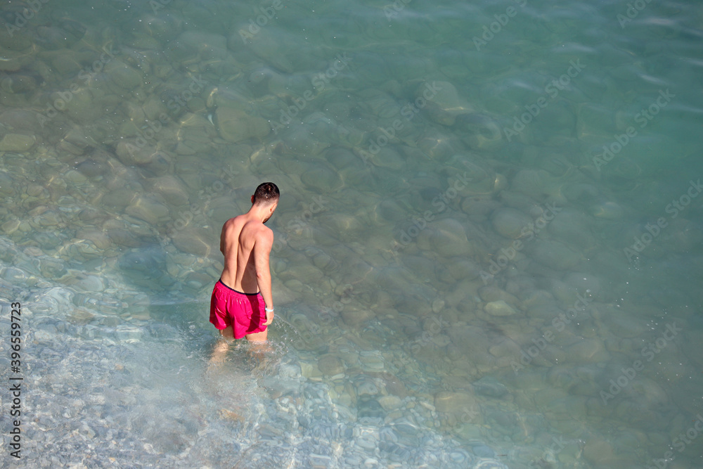 Man in red shorts going to swim in a sea. Aerial view to the pebble beach with transparent water, background for vacation and travel