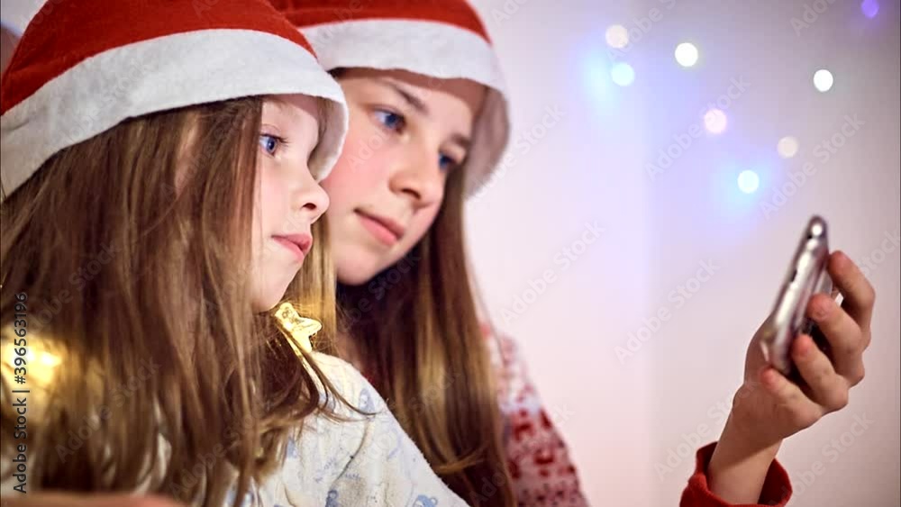 two sisters in Santa Claus hats watch cartoons on their smartphone on Christmas eve in a room with garlands, Christmas in quarantine two sisters in Santa Claus hats watch cartoons on their smartphone on Christmas eve in a room with garlands, Christmas in quarantine