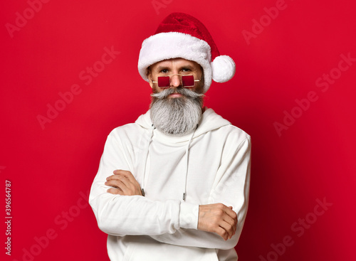 Winter fashion and sales. Portrait of a handsome senior man in a Santa hat, winking while standing against a red background. The hipster is holding glasses and looking at the camera. Place for text.
