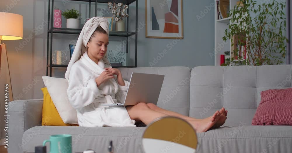 Close up of woman in bathrobe, towel sitting on sofa in living room speaking, having web conference using computer in home office. Makeup kits table in living room. Concept of modern technologies Close up of woman in bathrobe, towel sitting on sofa in living room speaking, having web conference using computer in home office. Makeup kits table in living room. Concept of modern technologies