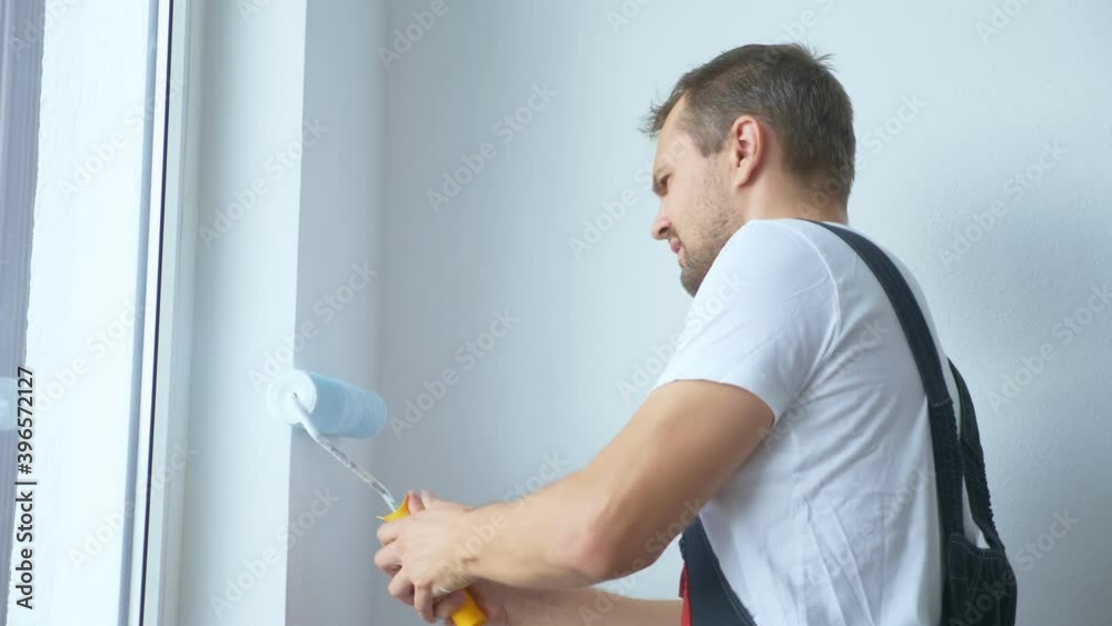 handsome man in work overalls paints the wall with a roller white paint. handsome man in work overalls paints the wall with a roller white paint.
