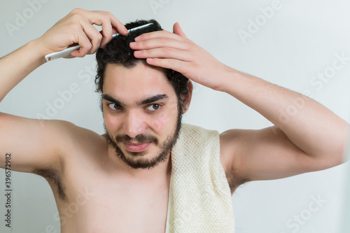caucasian young man with brown eyes and beard, combing his hair while standing in bathroom.