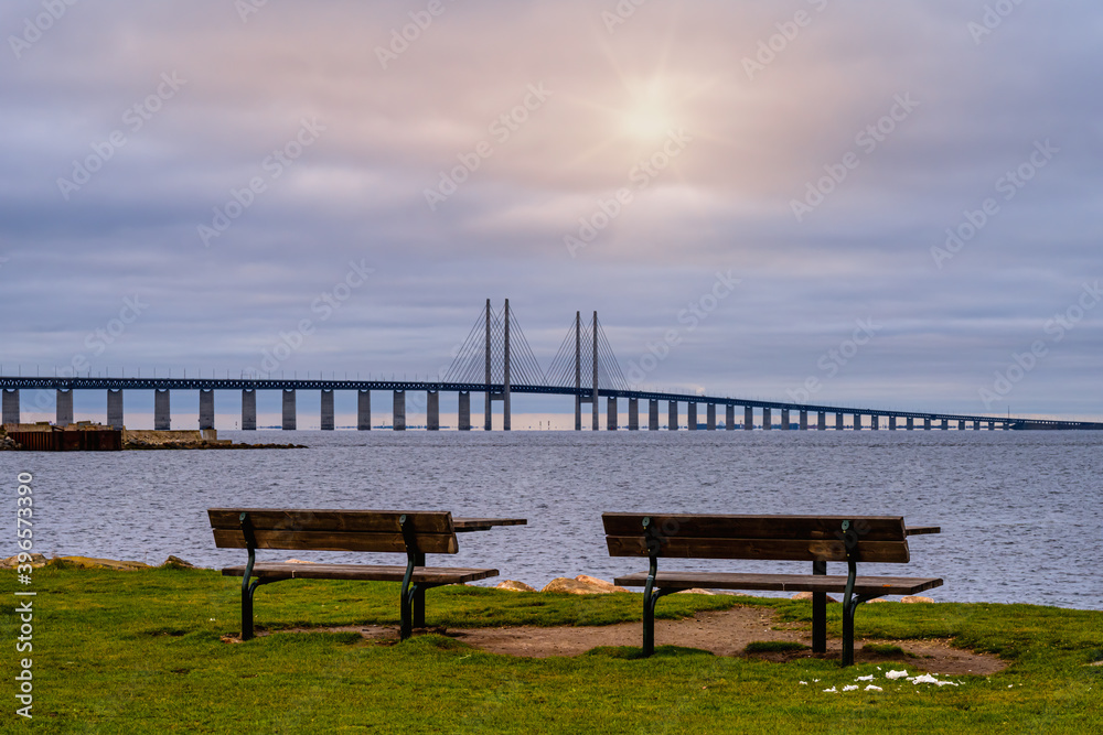 Two benches in front of a beautiful ocean view with a bridge. Blue ocean and a sunset sky in the background. Picture from Malmo, southern Sweden Two benches in front of a beautiful ocean view with a bridge. Blue ocean and a sunset sky in the background. Picture from Malmo, southern Sweden
