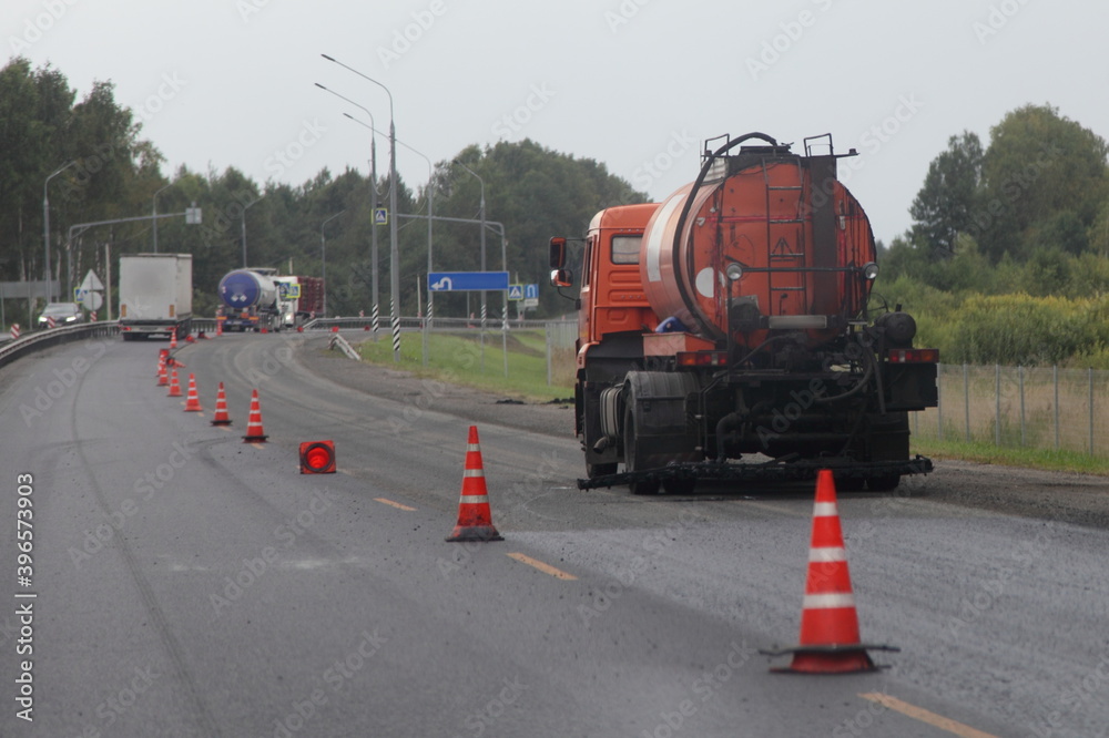 Orange bitumen truck pours liquid tar on the road behind the safety cones fencing, preparing to repair asphalt during road works at summer day