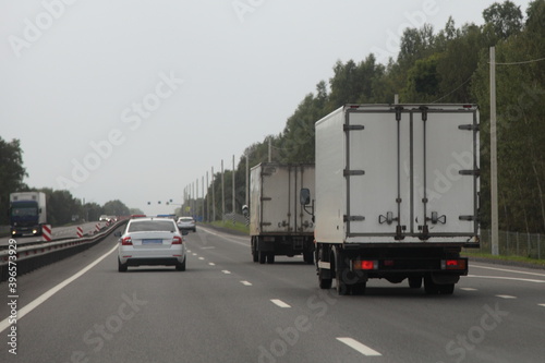 European trucks convoy with white van drive with police car on two lane suburban asphalted highway road at summer evening, rear view on forest and gray sky background, transportation logistics