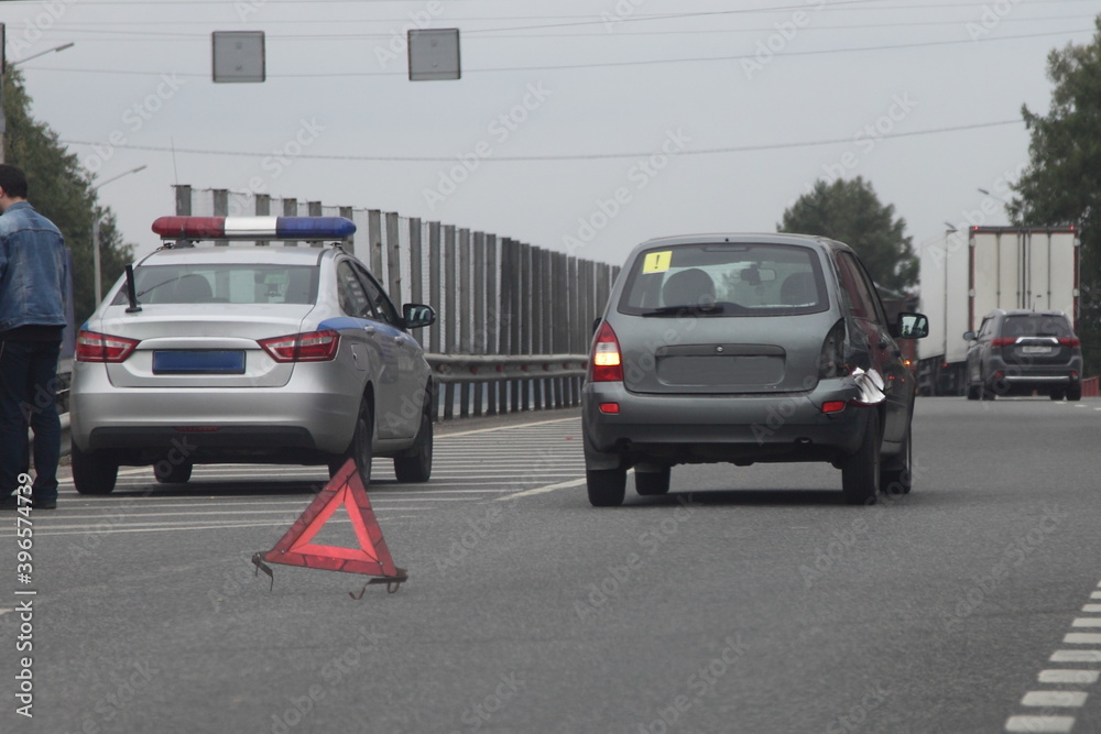 Road accident in Russia, compact broken car wreck wiyh damaged tail light and Russian police car on the background of emergency stop sign red triangle on suburban highway road at summer evening Road accident in Russia, compact broken car wreck wiyh damaged tail light and Russian police car on the background of emergency stop sign red triangle on suburban highway road at summer evening