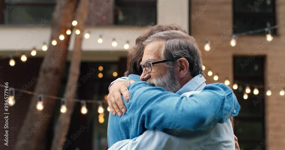 Cheerful happy Caucasian man meeting with old father and hugging at back yard of house. Family dinner outdoors on background. Senior dad with adult son in hugs. Generations. Two men embracing.