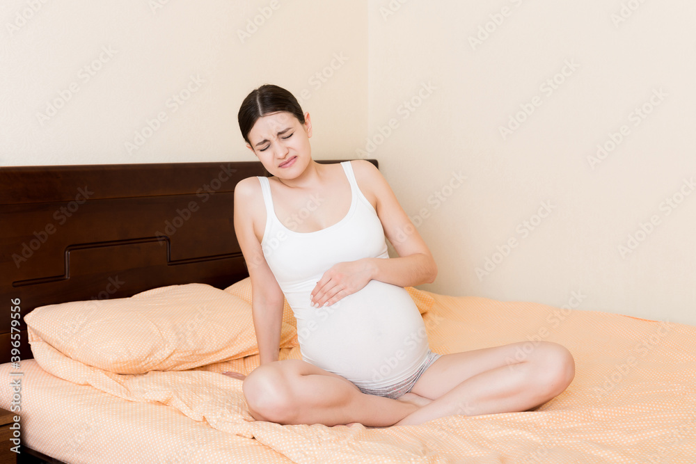 Young pretty pregnant woman, in pain, sitting on bed in bedroom, preparing to give a birth Young pretty pregnant woman, in pain, sitting on bed in bedroom, preparing to give a birth