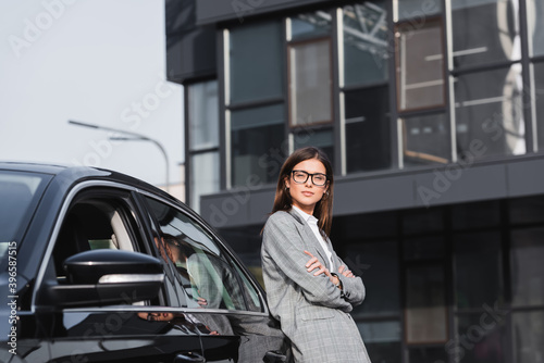  stylish businesswoman leaning on black car while standing with crossed arms